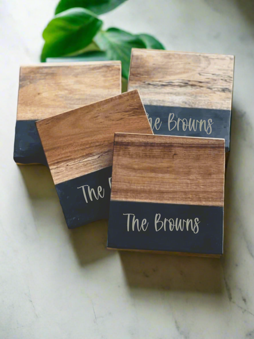 Four stacked acacia & slate square coasters on a white counter with a plant nearby