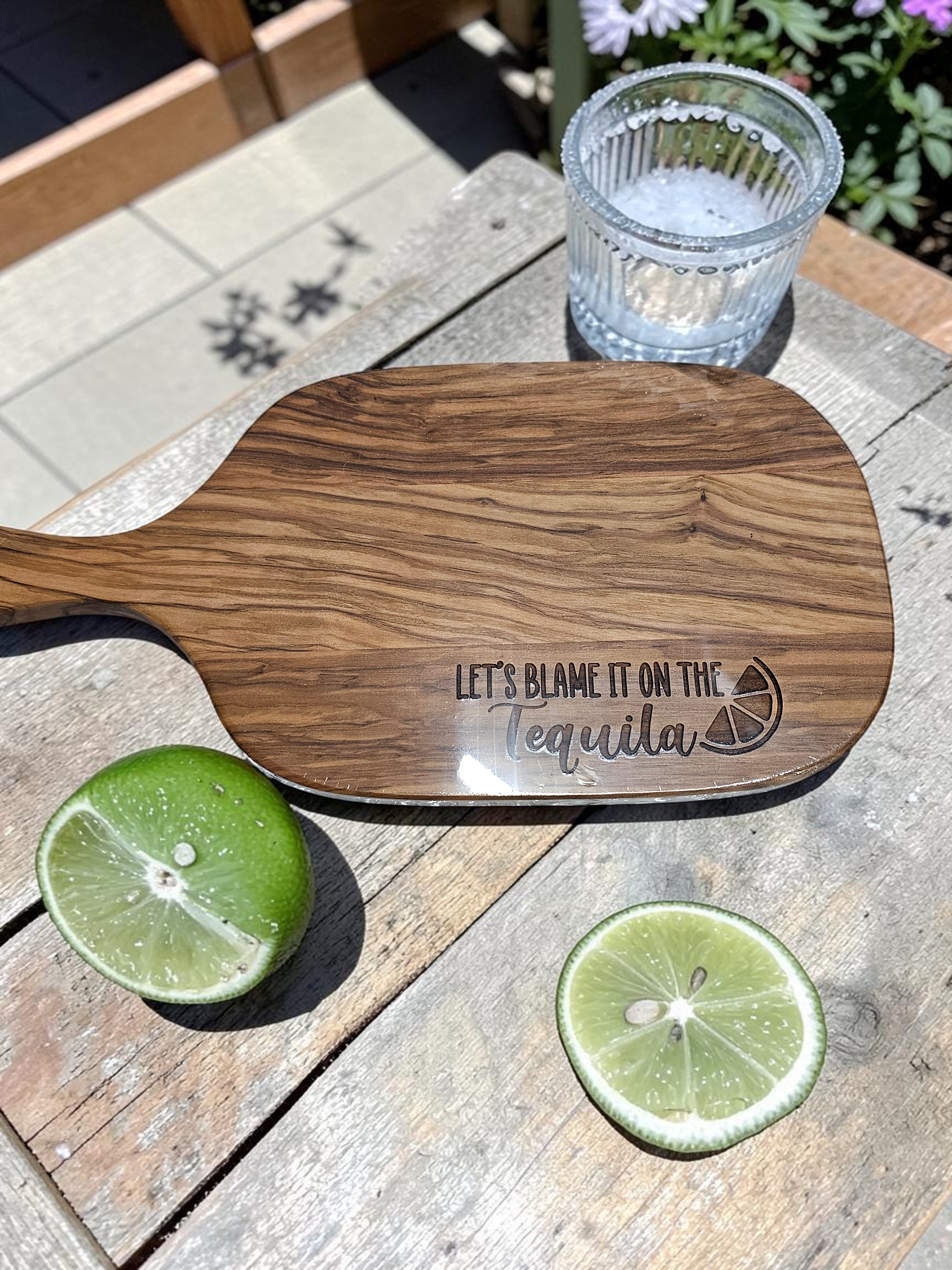 Wooden paddle with 'Let's Blame It on the Tequila' engraving on a wooden surface with limes and a glass.