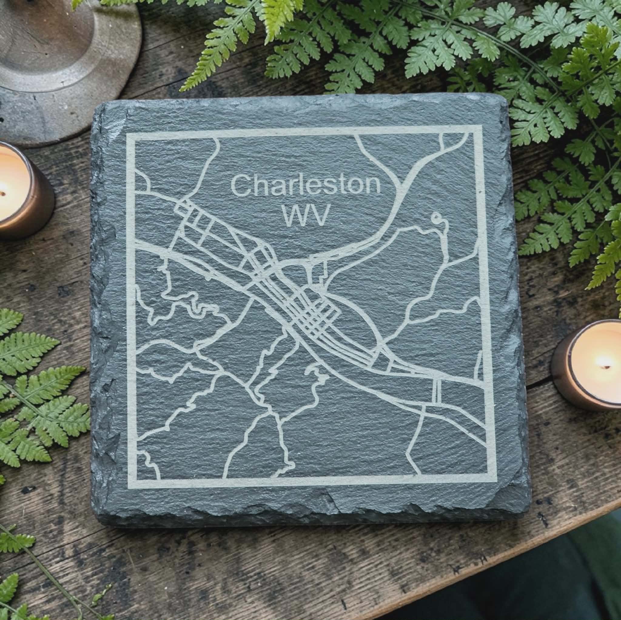Slate coaster engraved with a map of Charleston, WV, on a wooden background with greenery