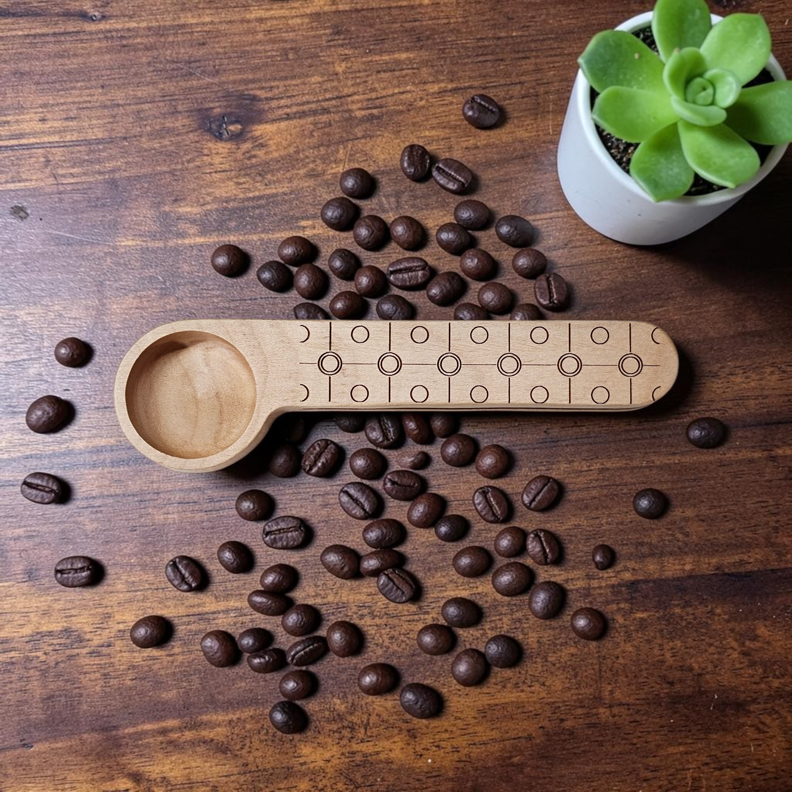 A light natural wood coffee scoop with engraved geometric patterns, displayed on a dark wooden tabletop surrounded by scattered coffee beans, with a small green succulent in a white pot in the upper right corner.