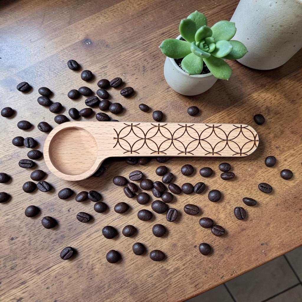 A small wooden coffee scoop with a laser-engraved geometric pattern on the handle, displayed on a wooden tabletop surrounded by scattered coffee beans, with a small potted succulent and a ceramic pot in the background.