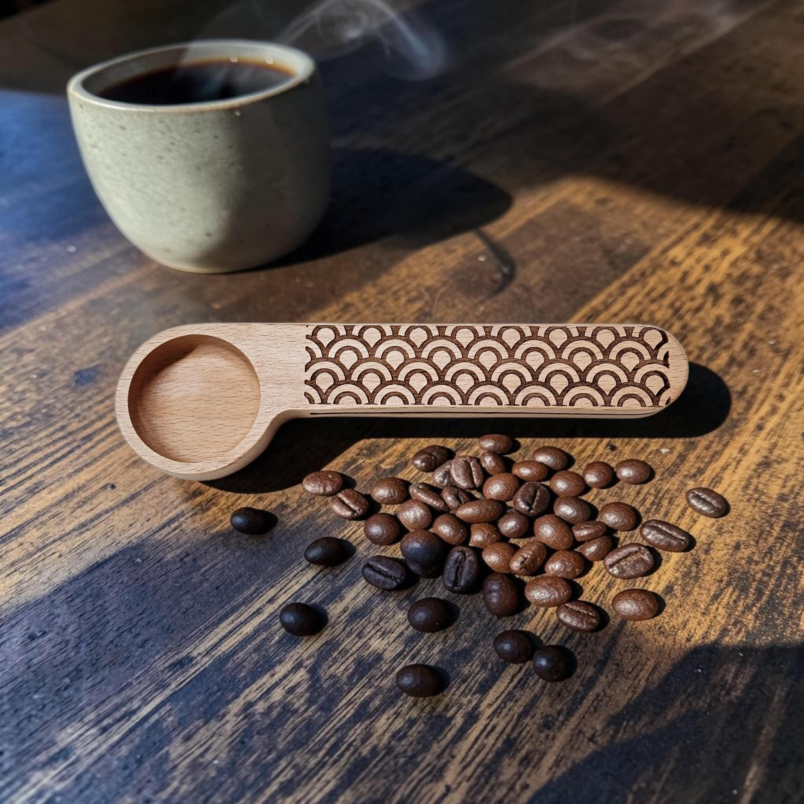A wooden coffee scoop with a laser-engraved wave pattern on the handle, displayed on a rustic wooden table next to roasted coffee beans and a cup of coffee in the background.