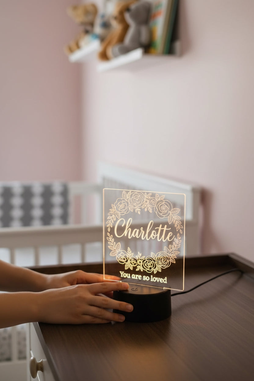 A personalized 3D acrylic LED night light with the name 'Charlotte' and 'You are so loved' engraved, shown being held by hands on a wooden table in a softly lit nursery.