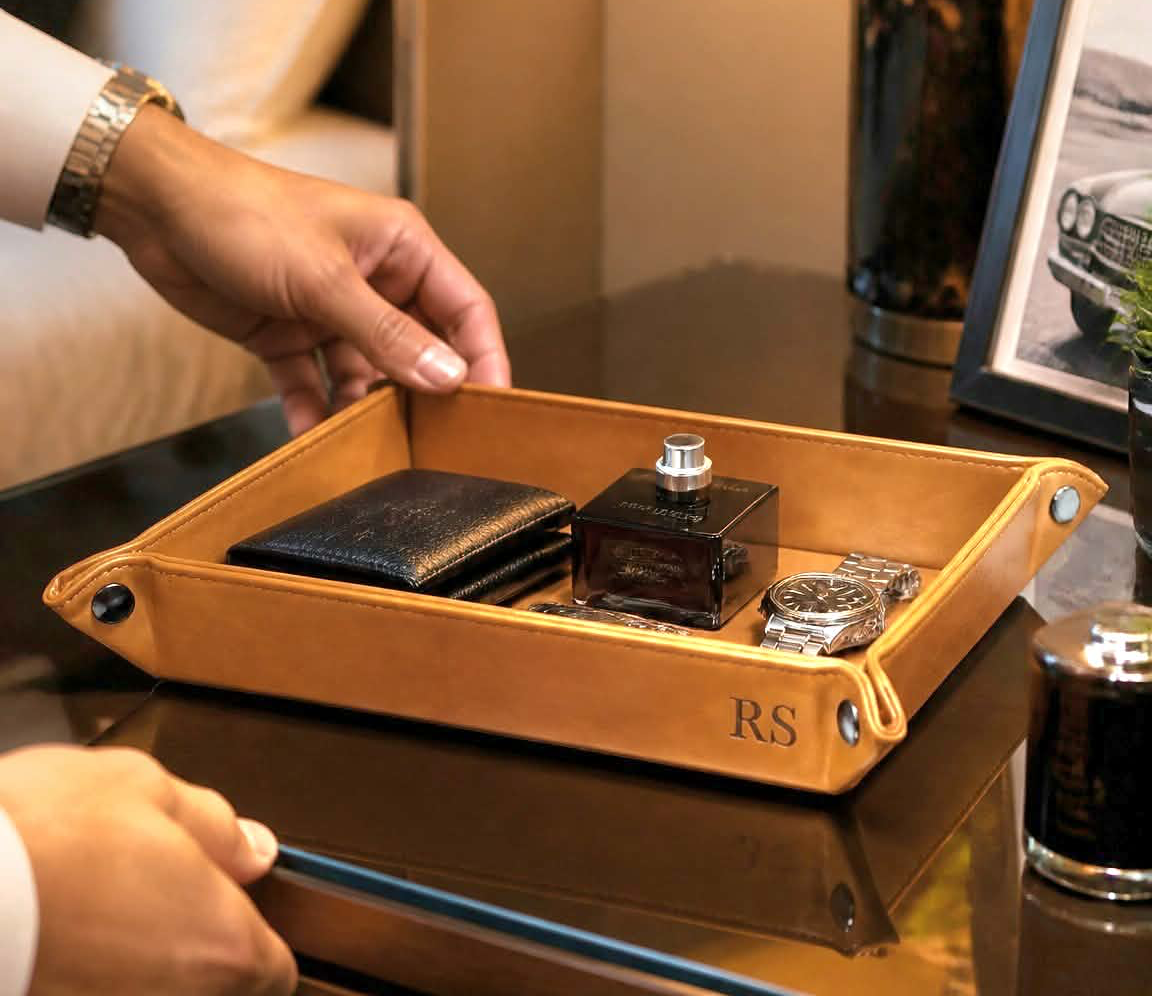 A close-up lifestyle shot of a person's hands placing a personalized light brown leather tray onto a dark wood bedside table. The tray features the initials 'RS' in gold on the bottom right corner and is styled with a black wallet, a perfume bottle, and a watch.