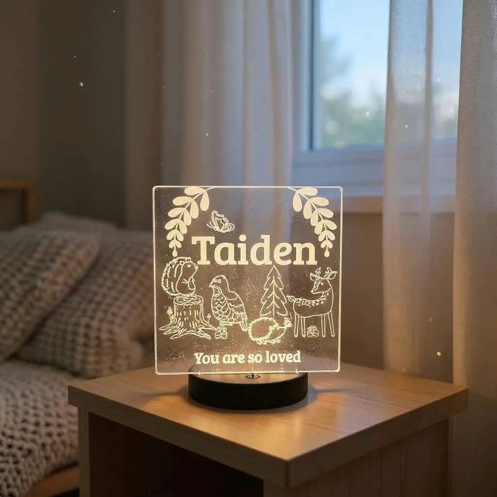 A personalized acrylic LED night light on a wooden bedside table, featuring the name 'Taiden' and woodland illustrations like a deer, tree, and bird, with the text 'You are so loved' in a cozy bedroom setting.