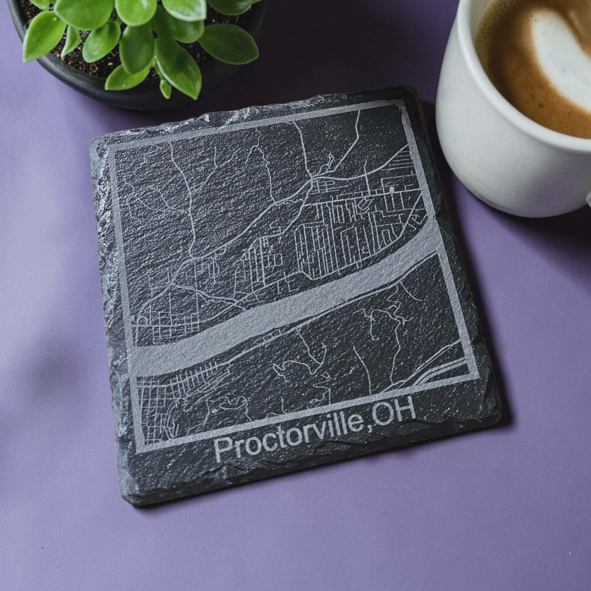 Slate coaster with engraved map and 'Proctorville, OH' text on a purple surface with a cup of coffee and plant.