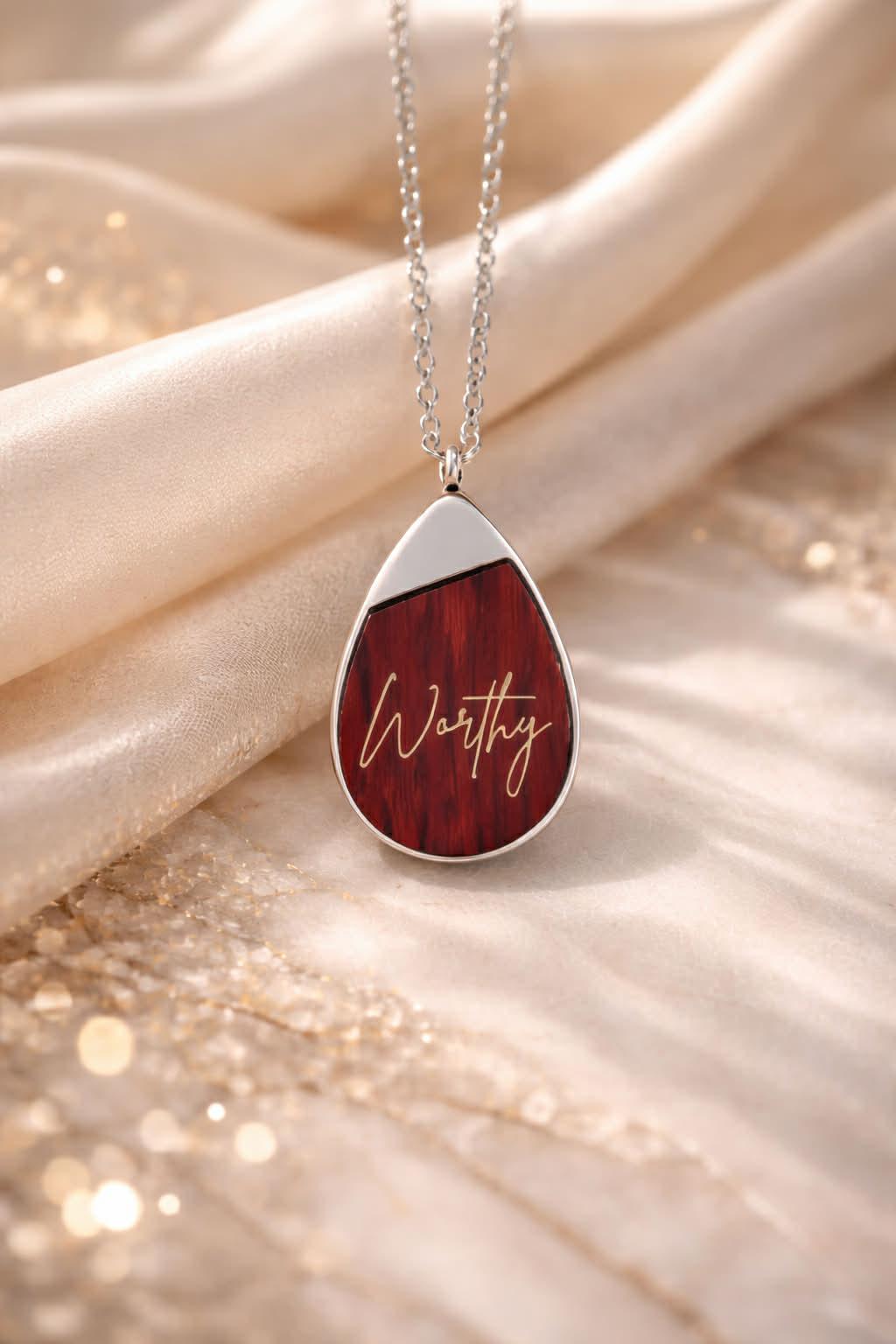Teardrop-shaped Rosewood pendant laser engraved with 'Worthy', hanging on a chain against a soft fabric background.