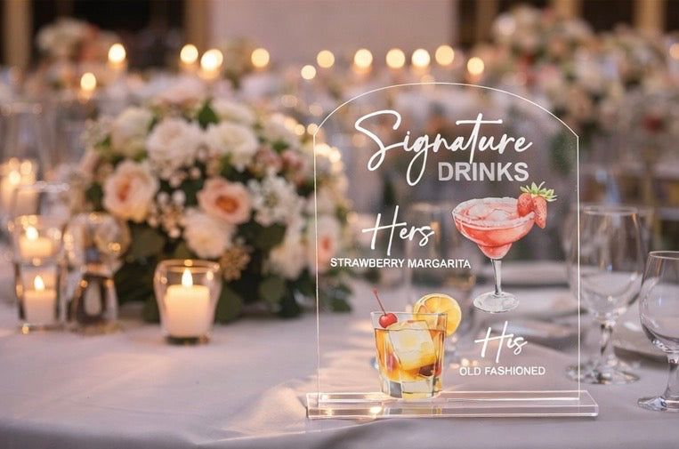 Acrylic sign for signature drinks at a wedding reception with floral decorations and candles.