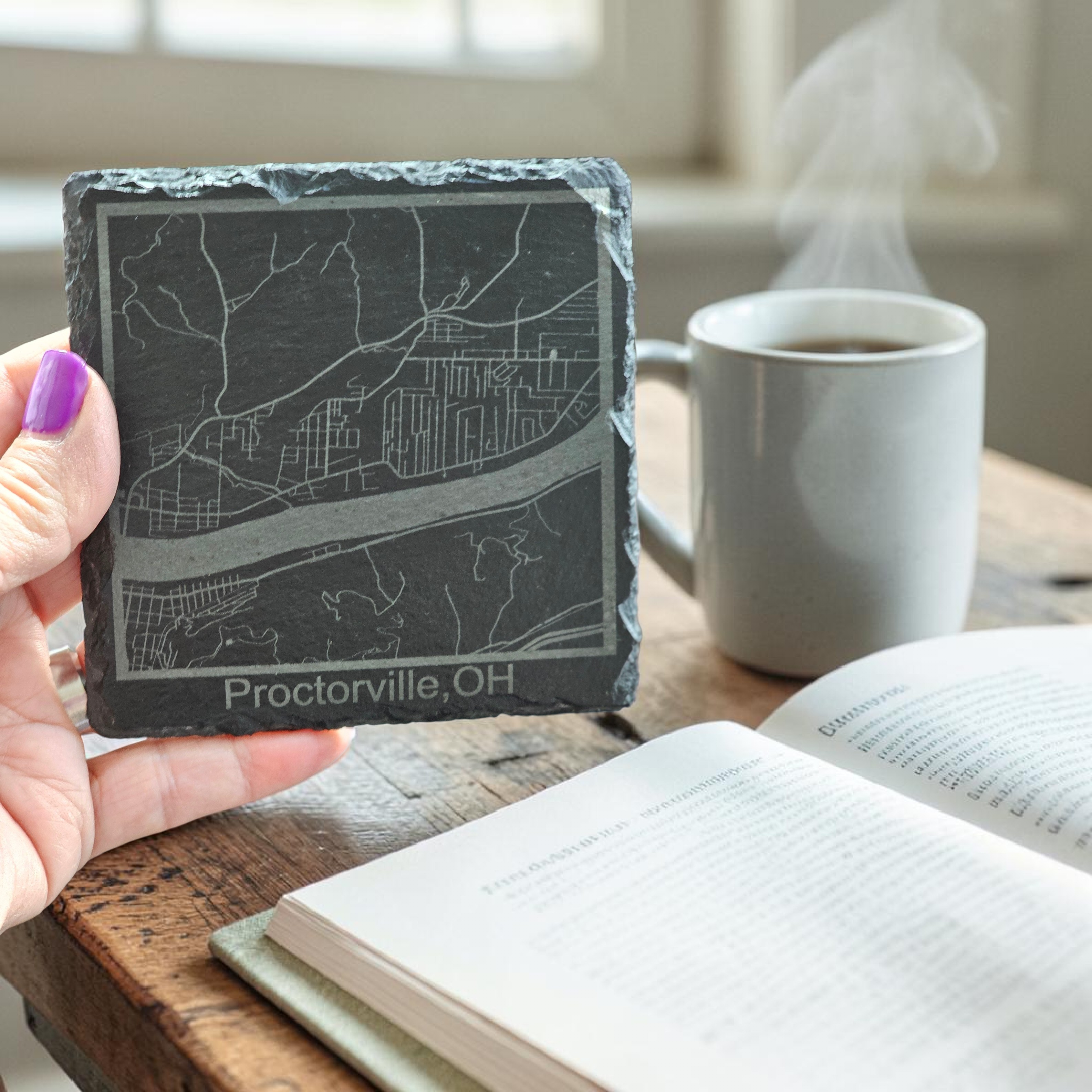 Slate coaster with engraved map and text held by a hand on a table with a book and coffee.